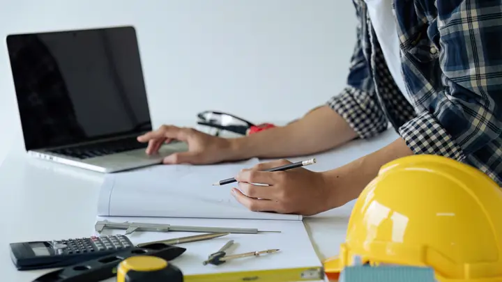 Hands working on a laptop and drawings with a calculator, tape measure and hard hat